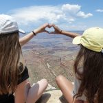 Kaksi tyttöä kuvattuna takaapäin muodostavat käsillään sydämen. Heidän edessään näkyy kanjoninäkymä.